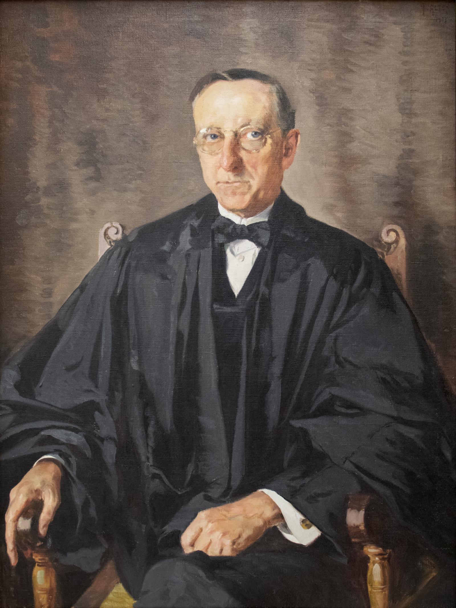 Portrait of a man with glasses seated in a wooden chair wearing black judicial robes.