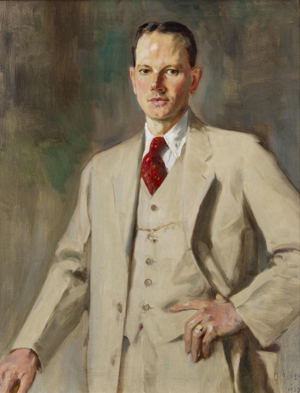 Portrait of man standing with one hand on left hip; wearing light-colored suit with red tie tucked into it.